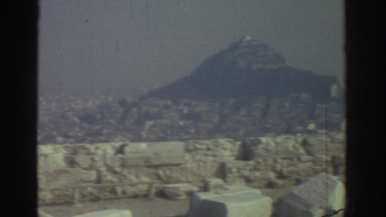 Cataloged 1976 8mm travel frame showing a hazy urban valley, distant hill, stone wall, and scattered ruins from an elevated viewpoint.