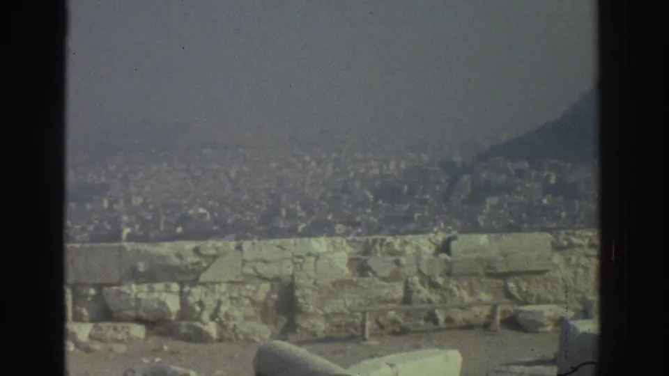 Hazy cataloged 1976 travel frame with a dense city below, low ancient stone wall, ruin fragments, and dark hillside at the edge.