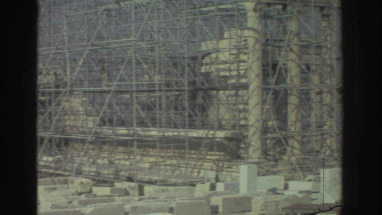 1970s 8mm frame of a scaffolded ancient stone structure, with masonry blocks in the foreground and dark frame edges.