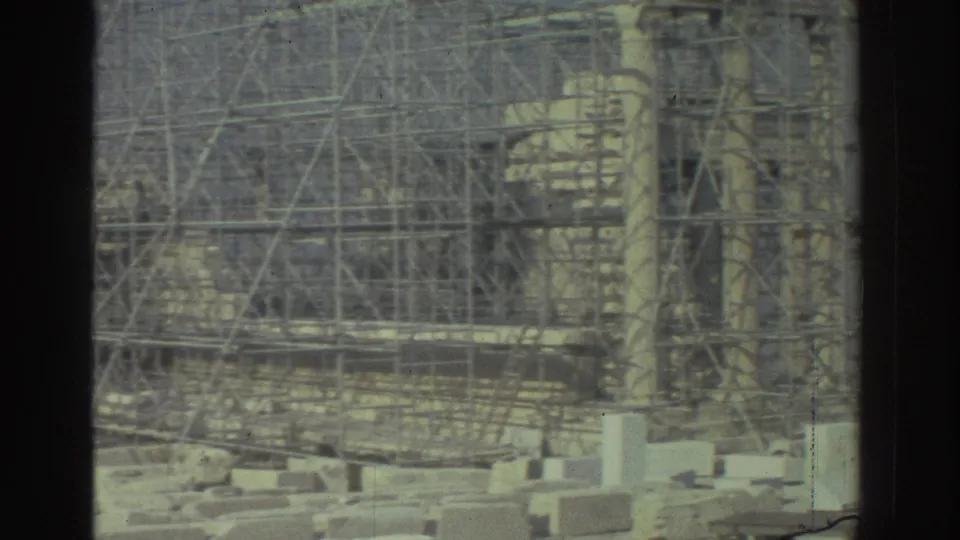 1970s 8mm frame of a scaffolded ancient stone structure, with masonry blocks in the foreground and dark frame edges.