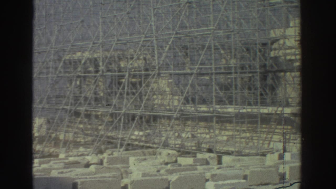 Muted 8mm view of scaffolded ancient stonework, seen through a dark vertical divider with loose blocks below.