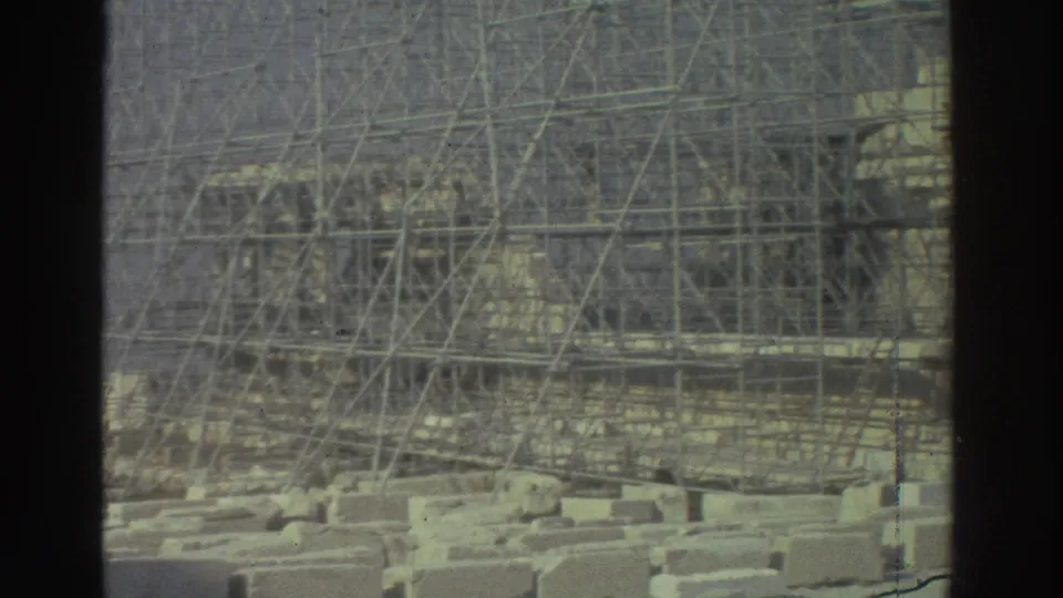 Muted 8mm view of scaffolded ancient stonework, seen through a dark vertical divider with loose blocks below.