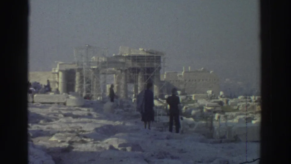 Wide 8mm frame of visitors near scattered stone blocks and a scaffolded ancient monument under a hazy sky.
