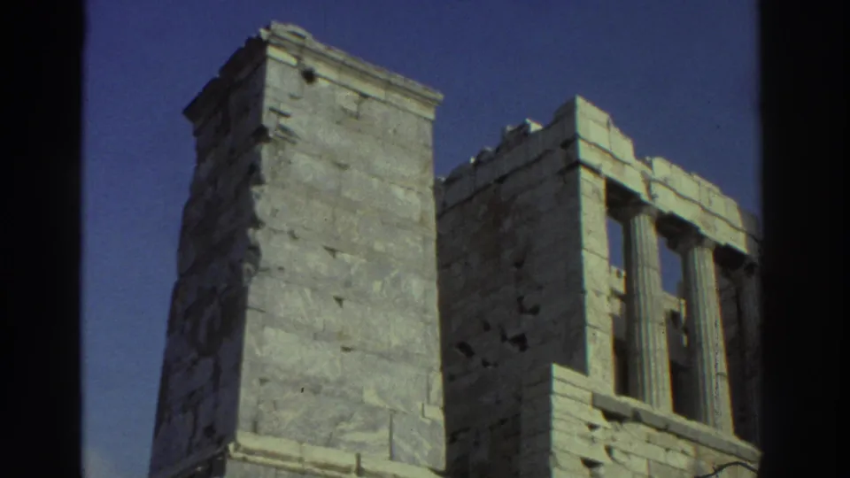 1970s 8mm travel frame showing a tall stone block structure and adjacent classical ruin elements beneath a clear blue sky.