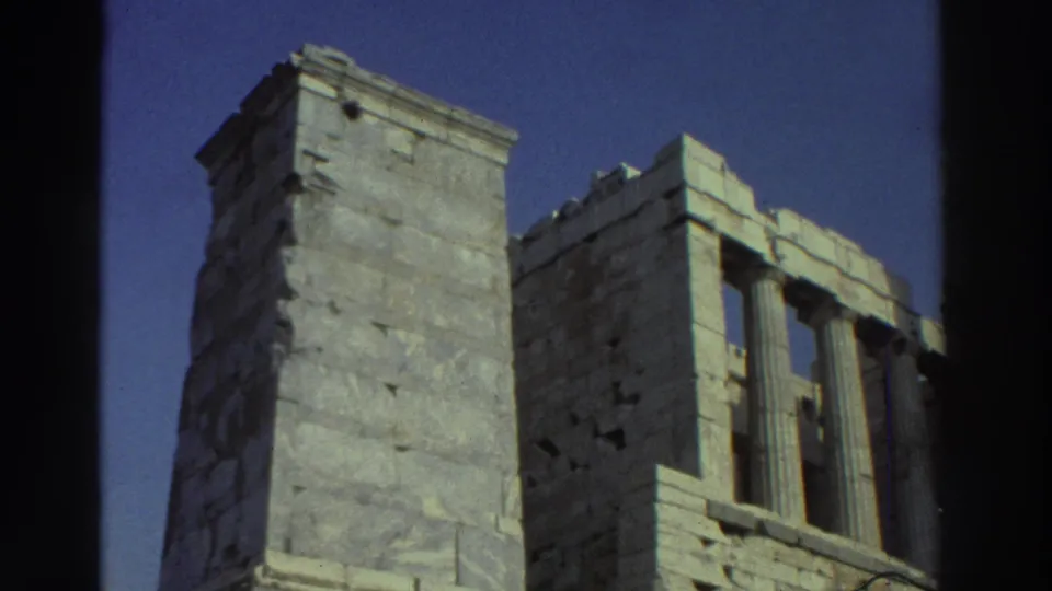 1970s 8mm frame of a weathered stone structure with adjoining classical masonry and deep blue sky, bordered by film edges.