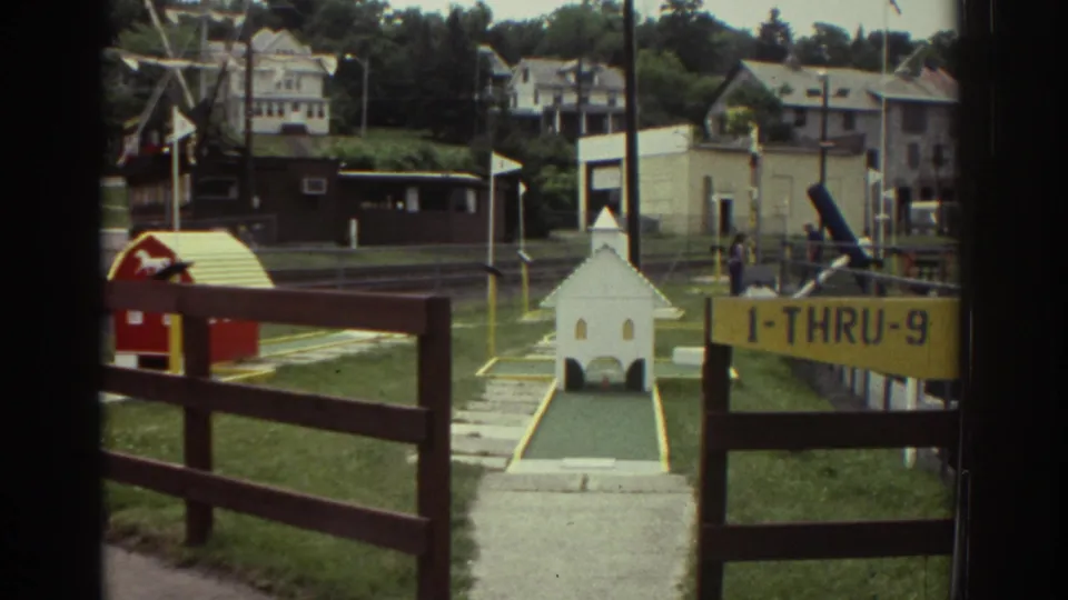 1976 Watkins Glen miniature golf course with a white church obstacle, red barn obstacle, yellow 1-THRU-9 sign, fencing, houses, and distant people.
