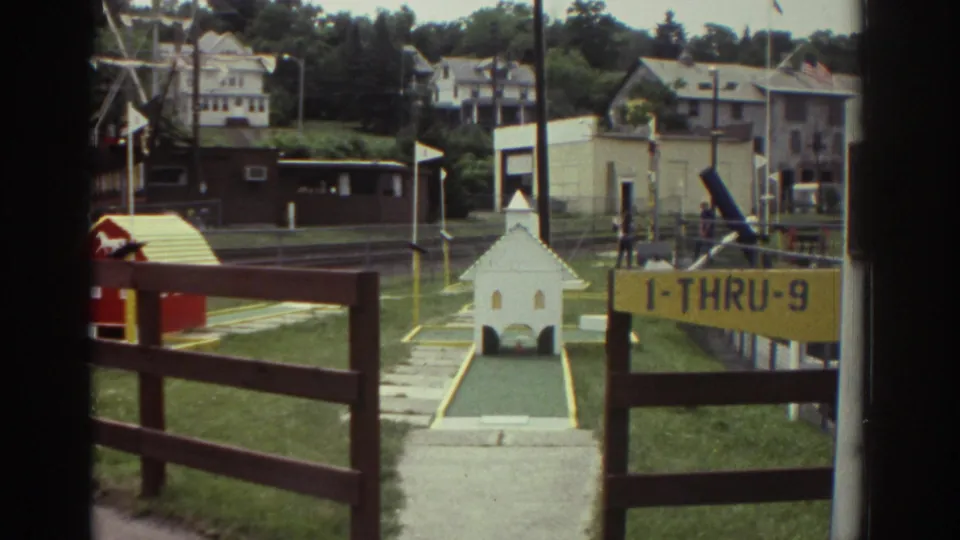 1976 Watkins Glen home-movie frame showing miniature golf holes with a white church obstacle, yellow 1-THRU-9 sign, fencing, houses, and distant visitors.