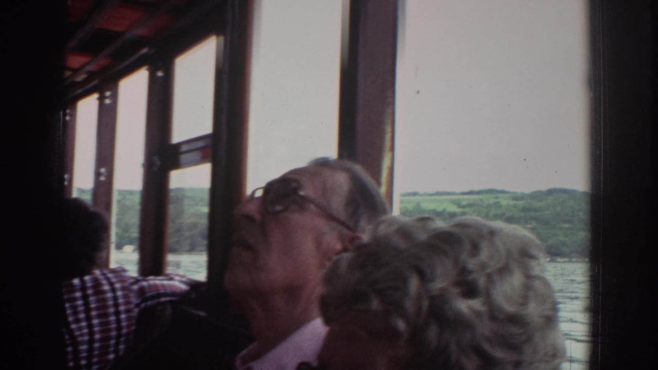 Older tourists aboard a 1976 Watkins Glen tour boat look out from the cabin toward lake water and wooded hills.