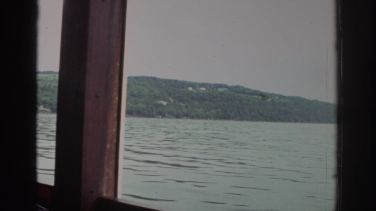 1976 Watkins Glen travel footage showing calm lake water and a wooded hillside framed by a wooden boat window.