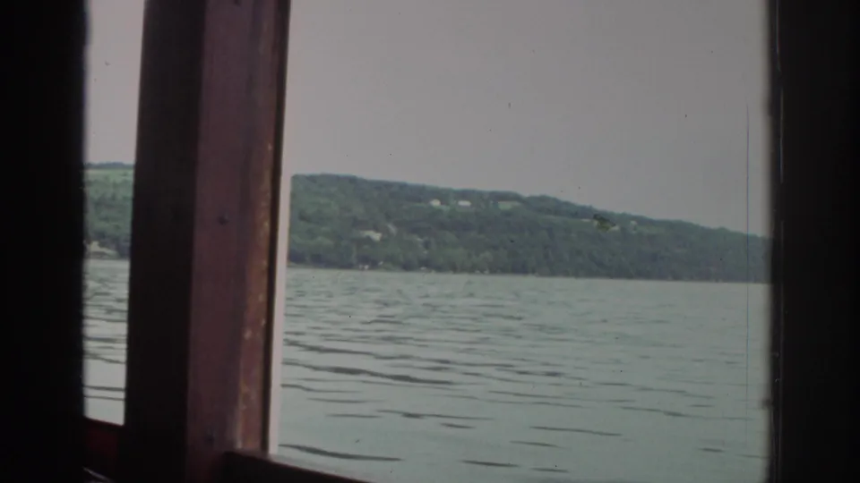 1976 Watkins Glen travel footage showing calm lake water and a wooded hillside framed by a wooden boat window.