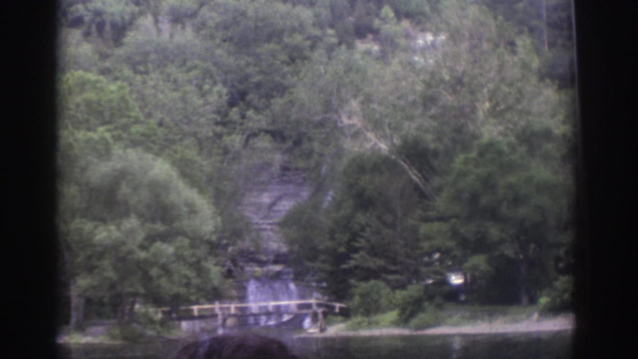 1976 Watkins Glen 8mm frame from a boat showing forested cliffs, calm water, a low shoreline bridge, and a partial passenger head in the foreground.