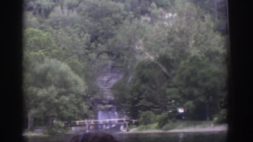 1976 Watkins Glen 8mm frame from a boat showing forested cliffs, calm water, a low shoreline bridge, and a partial passenger head in the foreground.