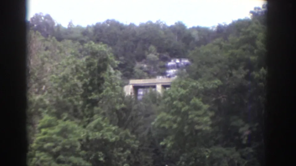 1976 Watkins Glen 8mm frame with a distant house or structure partly hidden by trees on a wooded hillside, seen from a moving boat.
