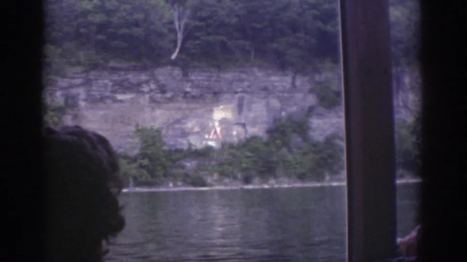 1976 Watkins Glen boat-view frame showing a rocky, tree-lined shore and cliff face, with a dark passenger silhouette and boat window post in front.