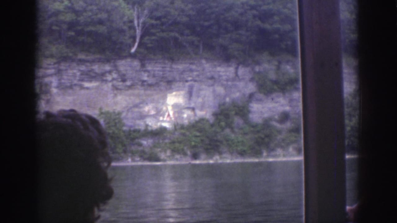 1976 Watkins Glen boat-window view with river water, rocky cliff, faint painted mural, trees, and a passenger silhouette in the foreground.