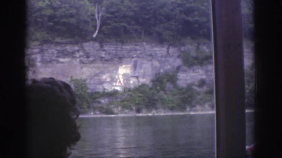 1976 Watkins Glen boat-window view with river water, rocky cliff, faint painted mural, trees, and a passenger silhouette in the foreground.