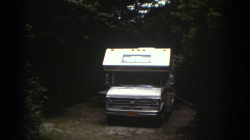 1976 Watkins Glen home-movie frame showing a white motorhome facing the camera in a shaded wooded clearing.