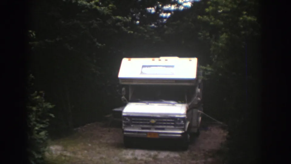 1970s home-movie view of a white motorhome parked on a small gravel clearing in the woods.