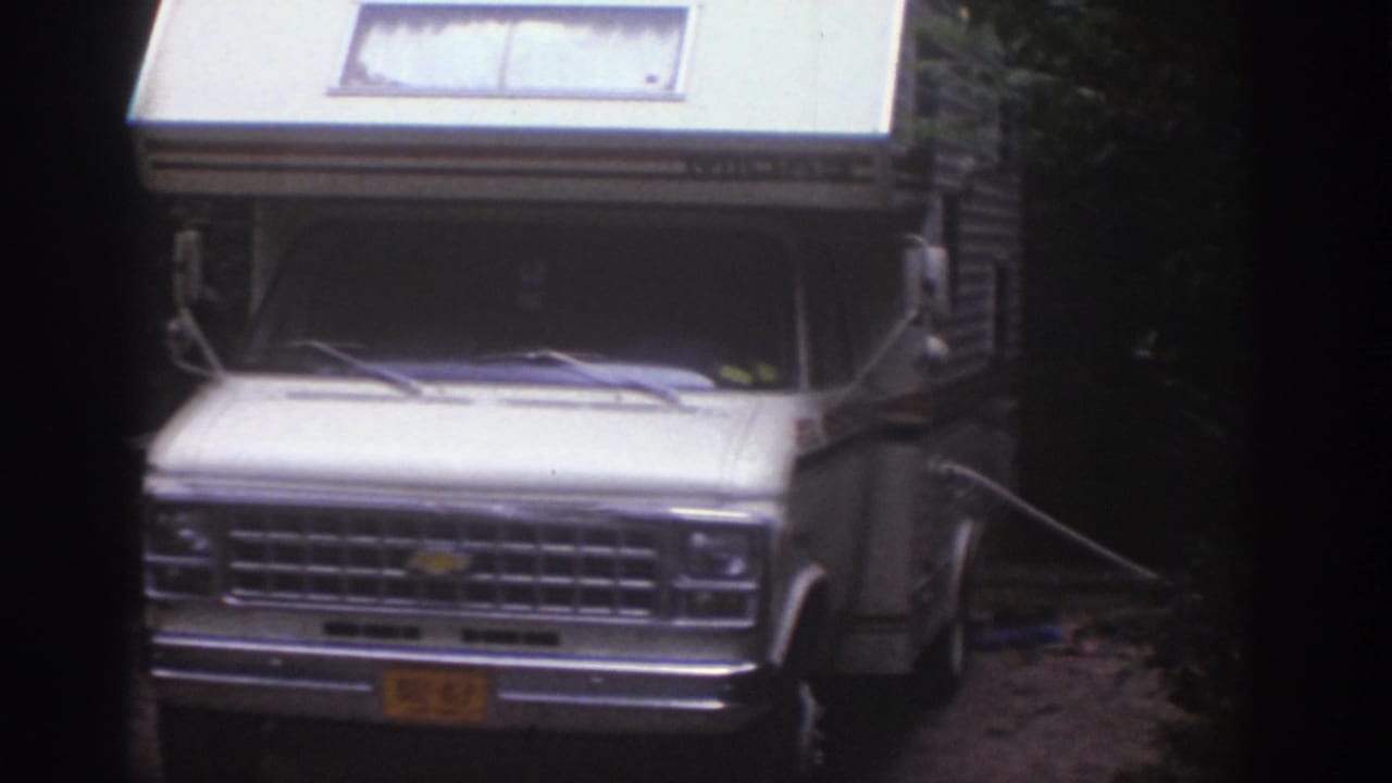 1976 Watkins Glen home-movie frame showing a white motorhome with raised roof parked on dirt near trees.
