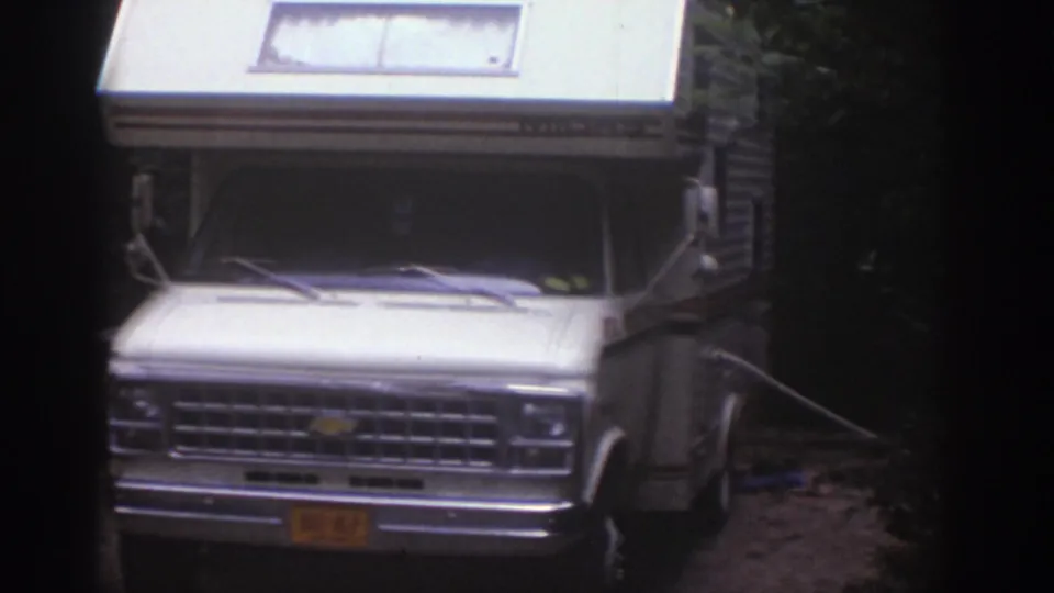 1976 Watkins Glen home-movie frame showing a white motorhome with raised roof parked on dirt near trees.