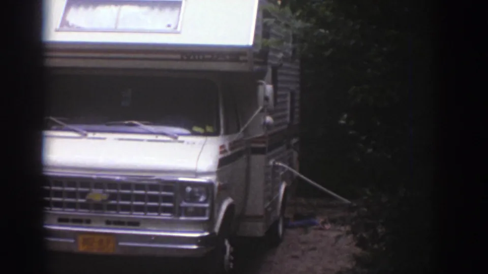 White Chevrolet-style motorhome with raised camper roof parked in a wooded Watkins Glen campsite in 1976 footage.