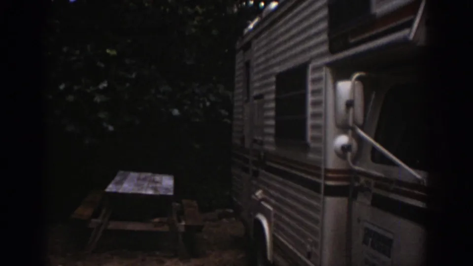 Side view of the parked motorhome at a wooded Watkins Glen campsite, with a picnic table visible nearby.