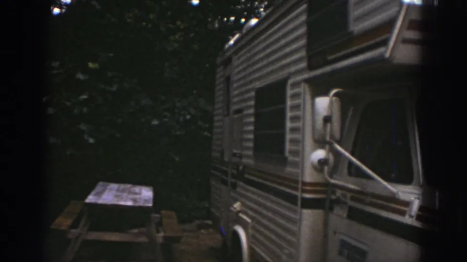 1976 Watkins Glen frame showing the motorhome side, side mirror, wooded background, and picnic table at the campsite.