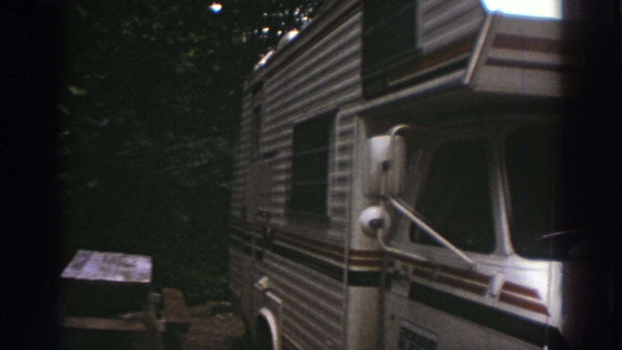 1976 Watkins Glen home-movie frame showing a white striped motorhome parked near a picnic table under dense trees.