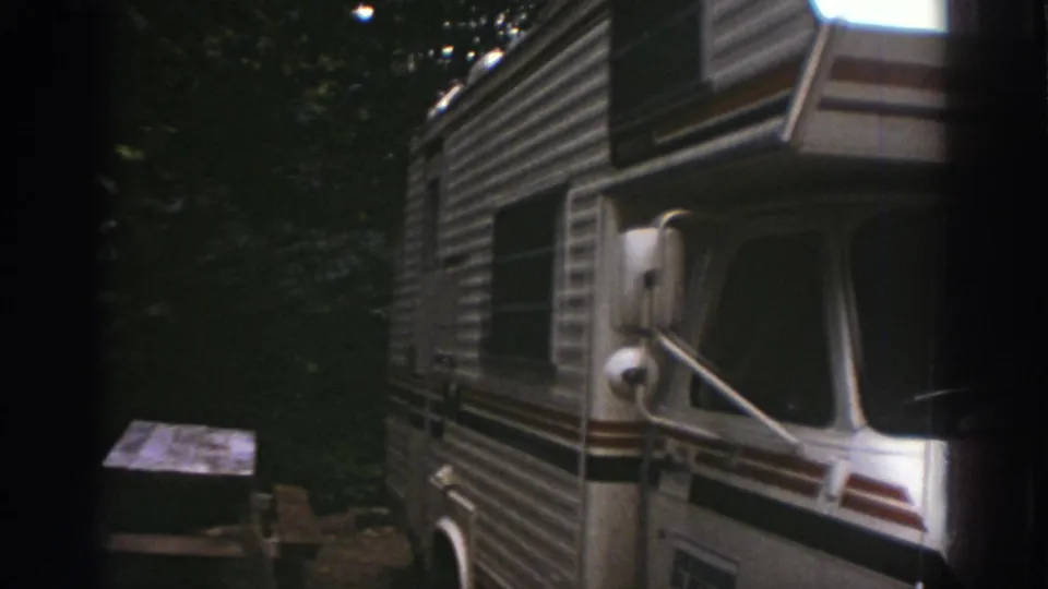 1976 Watkins Glen home-movie frame showing a white striped motorhome parked near a picnic table under dense trees.