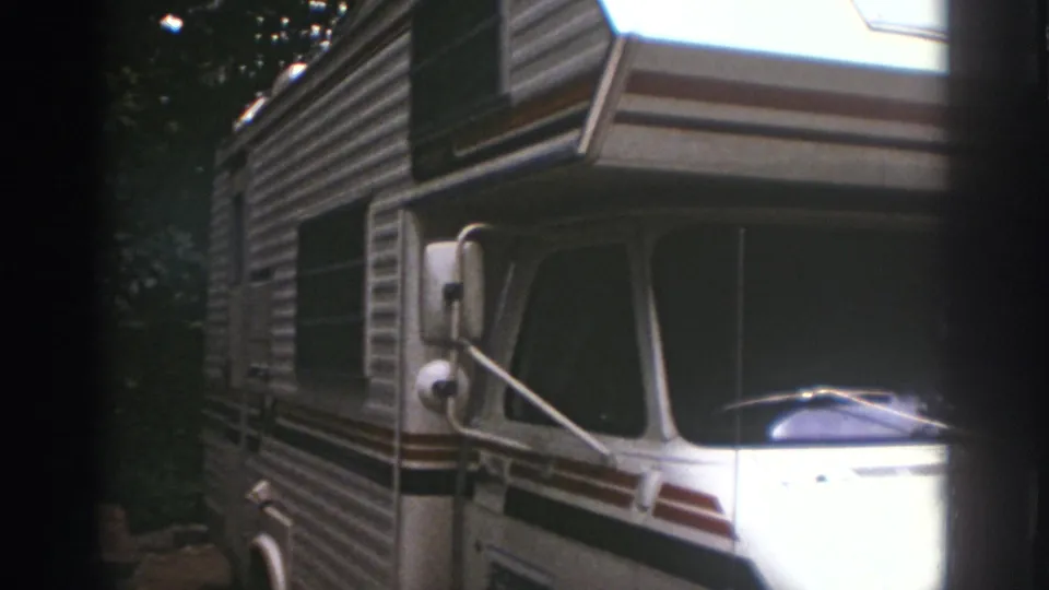 White 1970s motorhome with side mirror, windshield, striping, and shaded trees at the Watkins Glen campsite.