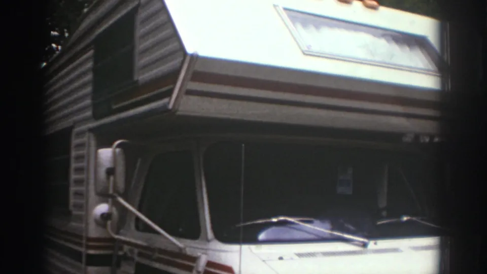 Final Watkins Glen campsite frame showing the motorhome cab, windshield wiper, mirror, stripes, and shaded wooded surroundings.