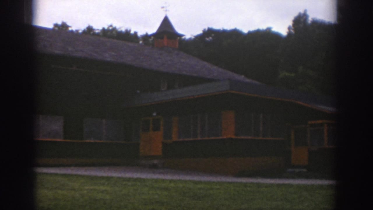 1976 Watkins Glen 8mm frame showing a low communal building with a small cupola, orange doors, grass, walkway, and wooded backdrop.