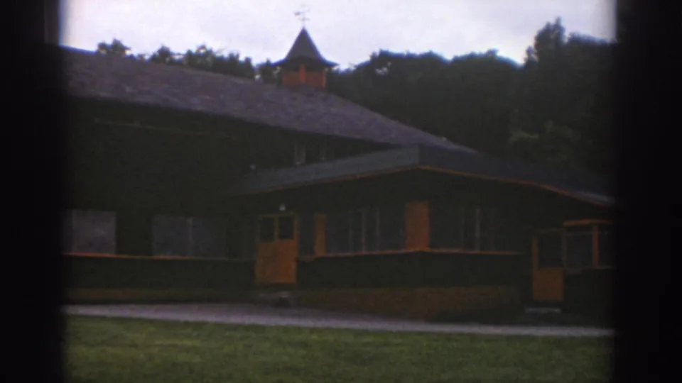 1976 Watkins Glen 8mm frame showing a low communal building with a small cupola, orange doors, grass, walkway, and wooded backdrop.