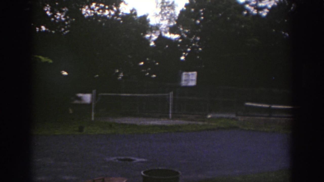 1976 Watkins Glen 8mm frame of an empty outdoor court area behind fencing, with a volleyball net, posted sign, and trees beyond.