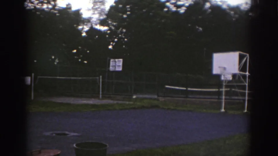 1976 Watkins Glen 8mm frame showing an empty sports court with volleyball net, basketball backboard, chain-link fence, and wooded edge.