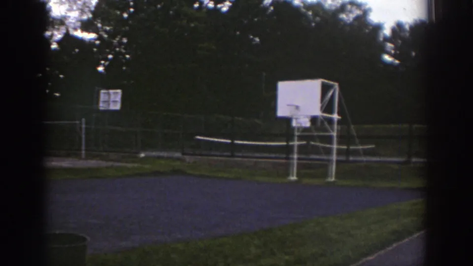 1976 Watkins Glen 8mm frame of an empty fenced recreation court with a white basketball backboard, volleyball net, trees, and grass foreground.