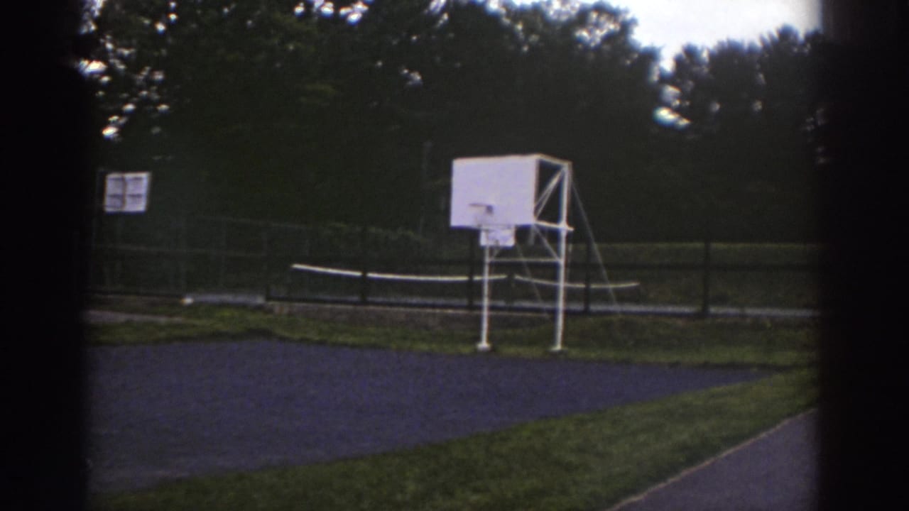 1976 Watkins Glen home-movie frame showing a quiet outdoor basketball court beside trees and fencing.