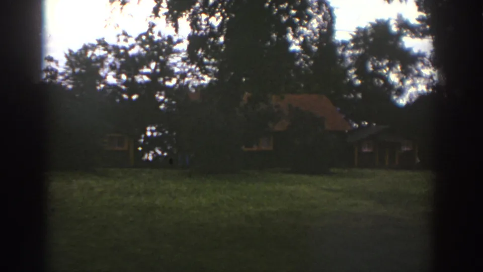 1976 Watkins Glen home-movie frame of a rustic cabin seen across a grassy wooded area.