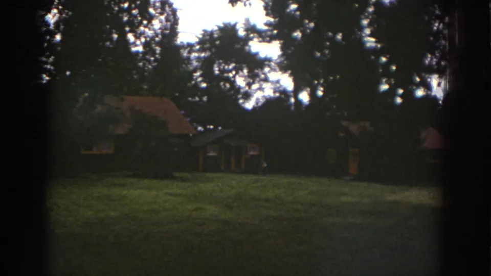 1976 Watkins Glen home-movie frame with the cabin and grassy foreground under heavy tree shadow.