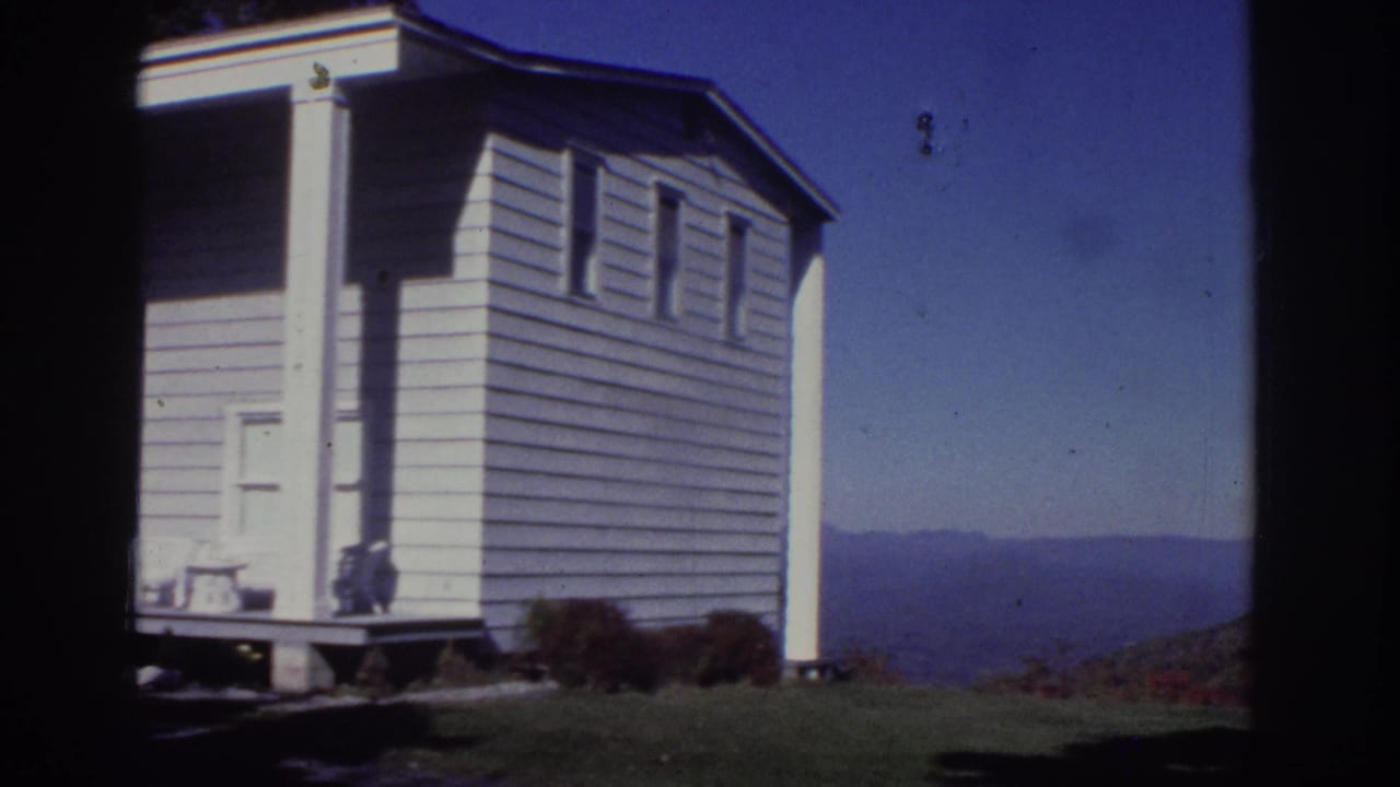1976 Watkins Glen home-movie frame showing a small white cottage side wall, lawn, shrubs, and distant hills under clear blue sky.