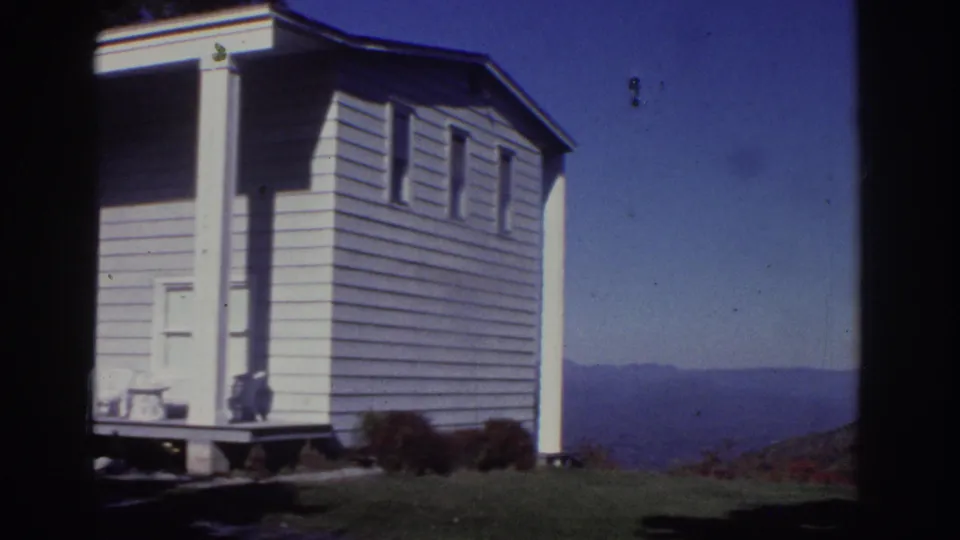 1976 Watkins Glen home-movie frame showing a small white cottage side wall, lawn, shrubs, and distant hills under clear blue sky.