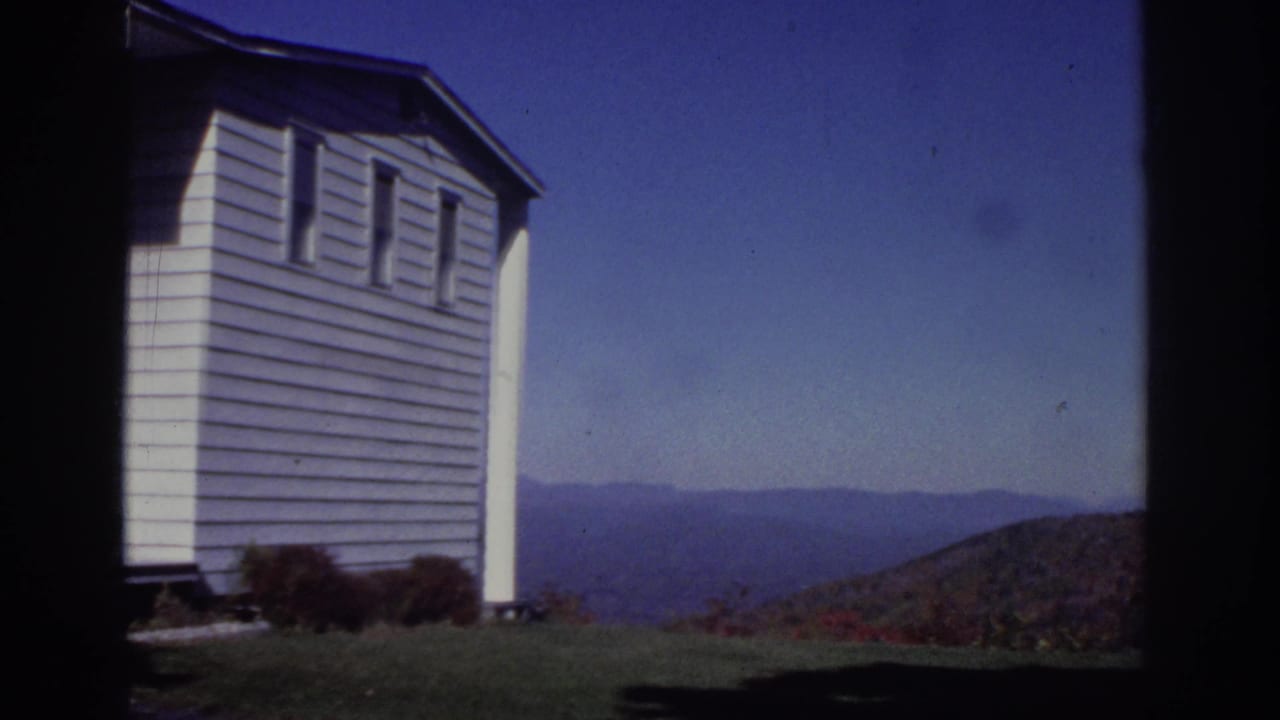 Side view of a white cottage with lawn, shrubs, blue sky, and distant hills.