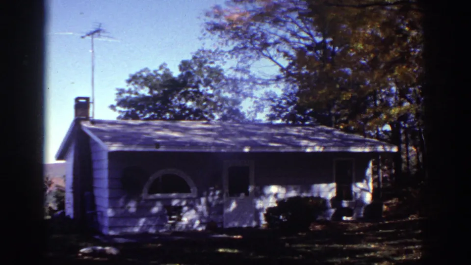 1976 Watkins Glen home-movie view of a white cottage front with porch, rooftop antenna, trees, and shaded grass.