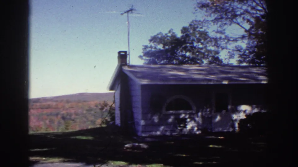 1976 Watkins Glen frame showing a white cottage front, roof antenna, trees, lawn, and autumn-toned hills in the distance.