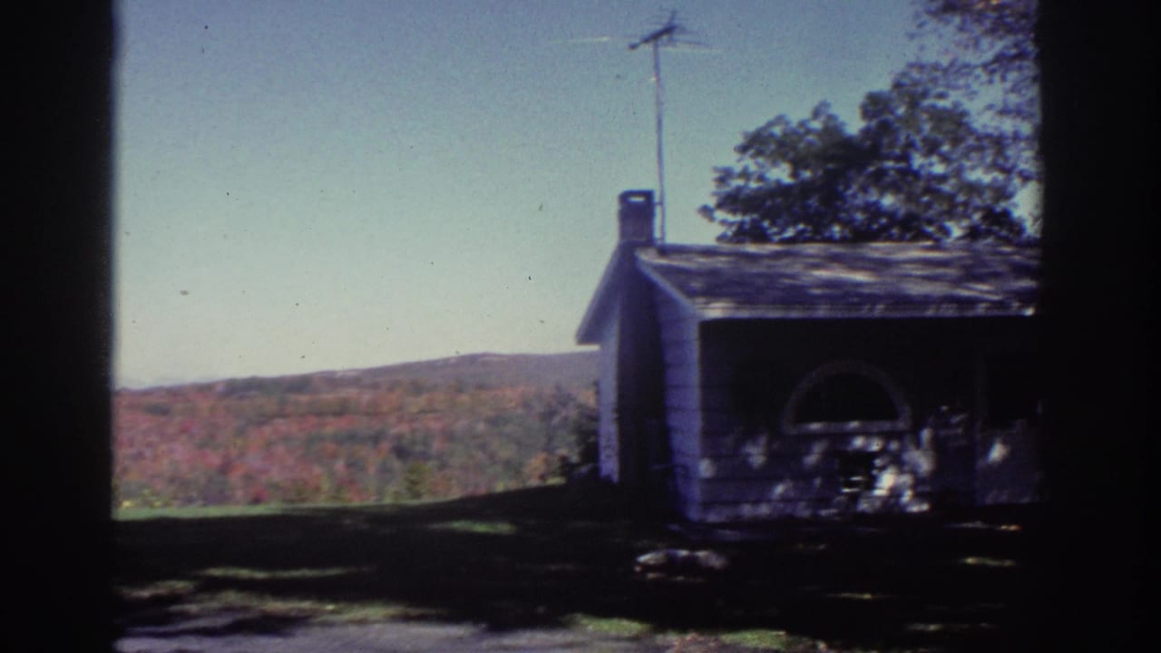 1976 Watkins Glen home-movie frame showing a white rural house, chimney, lawn, trees, and autumn hills beyond a dark window frame.