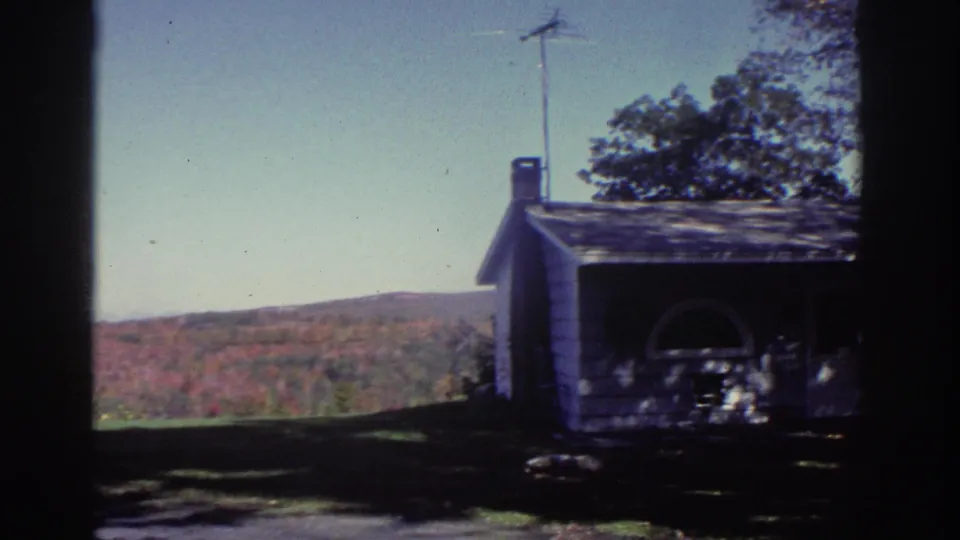 1976 Watkins Glen home-movie frame showing a white rural house, chimney, lawn, trees, and autumn hills beyond a dark window frame.