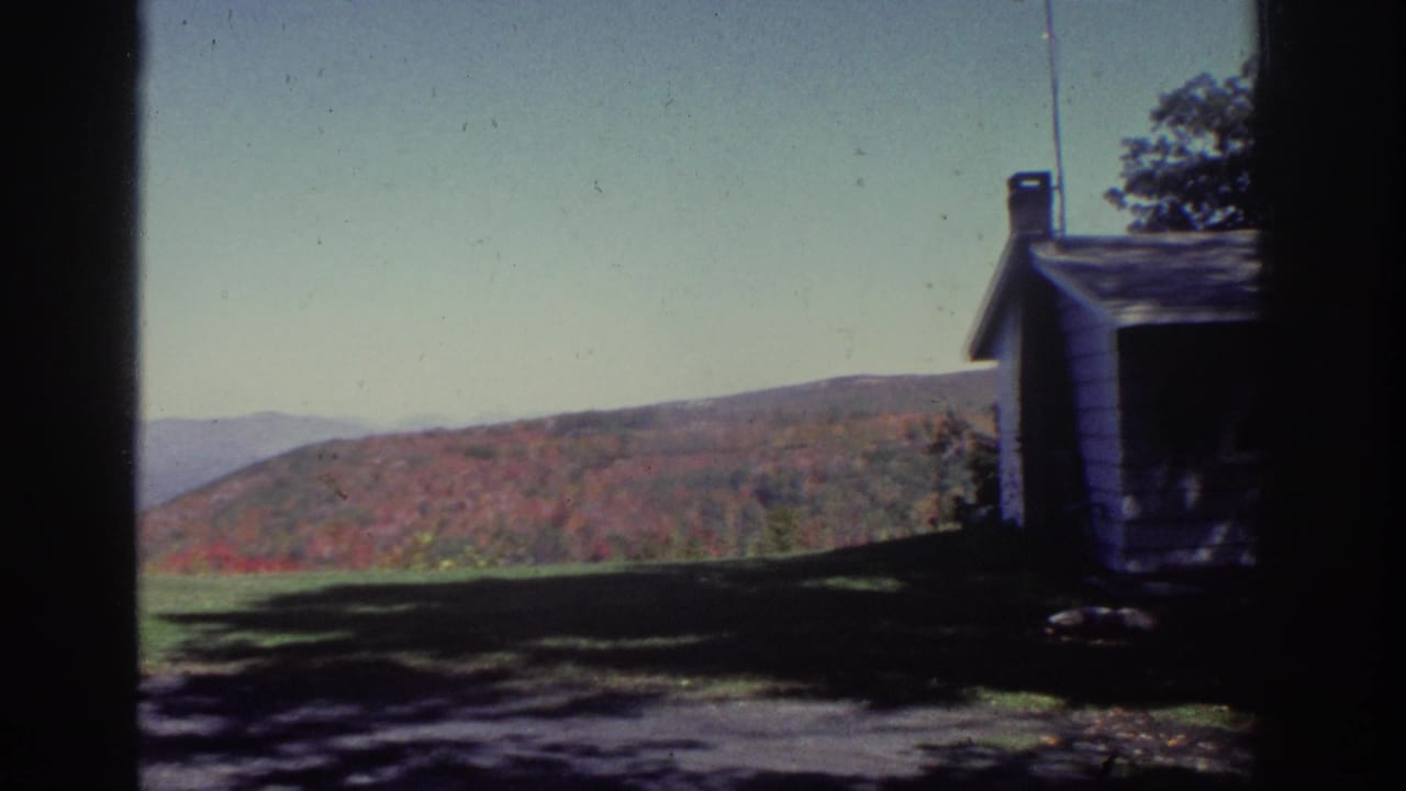 White house at the edge of a lawn with wooded autumn hills behind it.