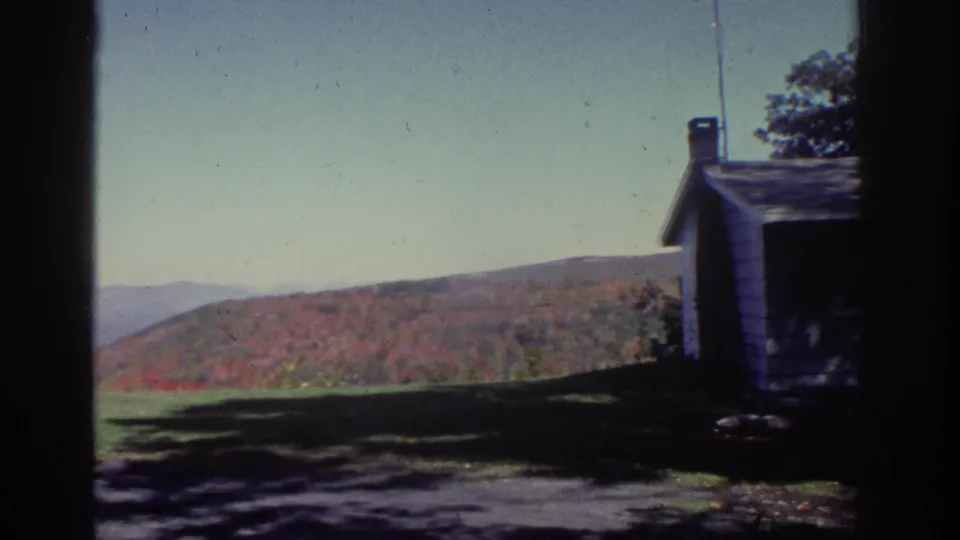 1976 Watkins Glen footage shows a rustic white house and chimney beside a lawn, with red and orange hills under a clear sky.