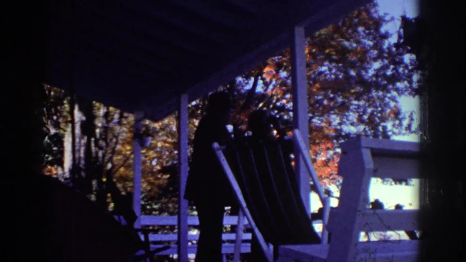 1976 Watkins Glen porch view with a silhouetted adult near a lounge chair, white railings, porch posts, and autumn trees beyond.
