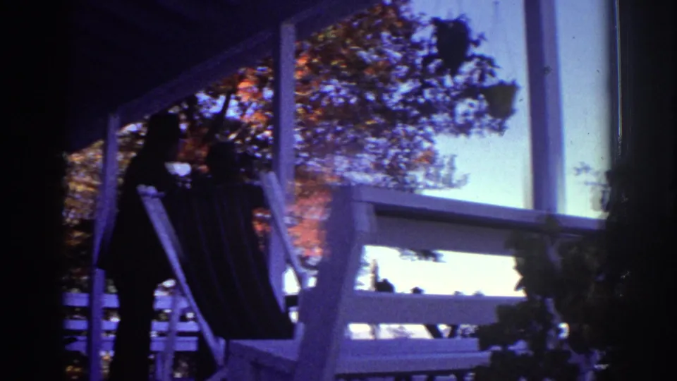 1976 Watkins Glen home-movie frame showing a shadowed porch, silhouetted adult, lounge chair, white railings, and bright autumn foliage.